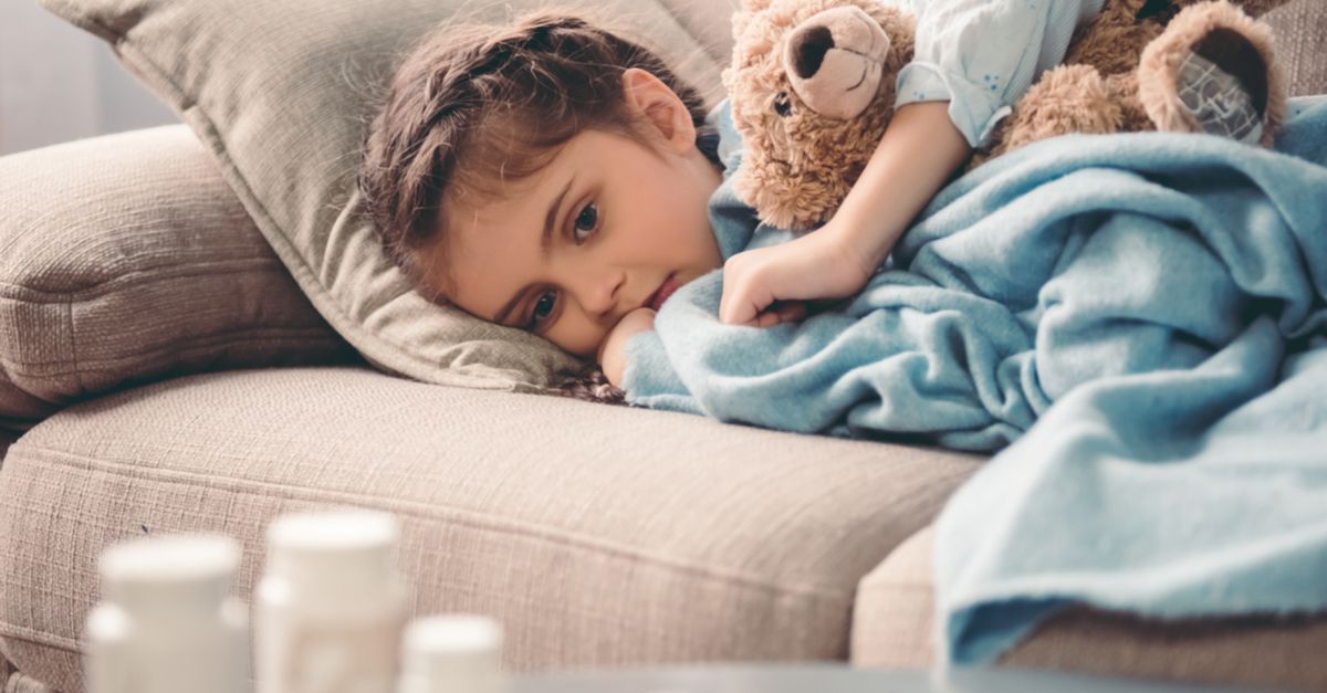Child laying on couch covered by blanket hugging teddy bear with pill bottles in the foreground