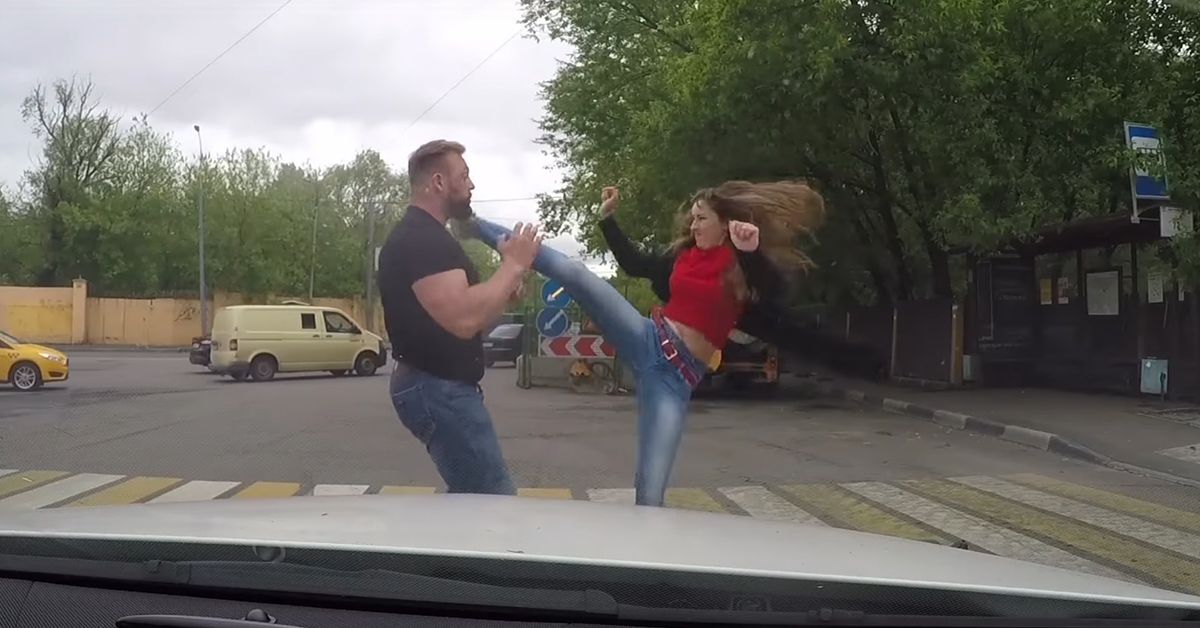 Still from a video showing a woman delivering a roundhouse kick to a man in front of a car