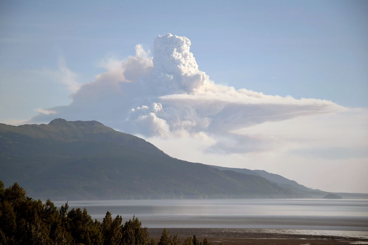 White smoke drifts from a mountain area. A lake area is below the mountains, near a forest area.