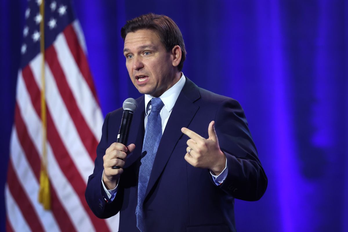 A white man speaking in a microphone stands near a blue backdrop with an American flag next to it.