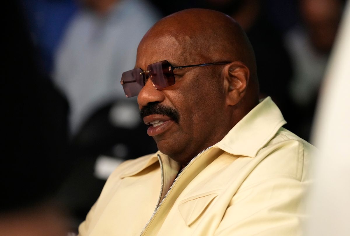 A Black man wearing sunglasses and a yellow shirt is sitting down. 
