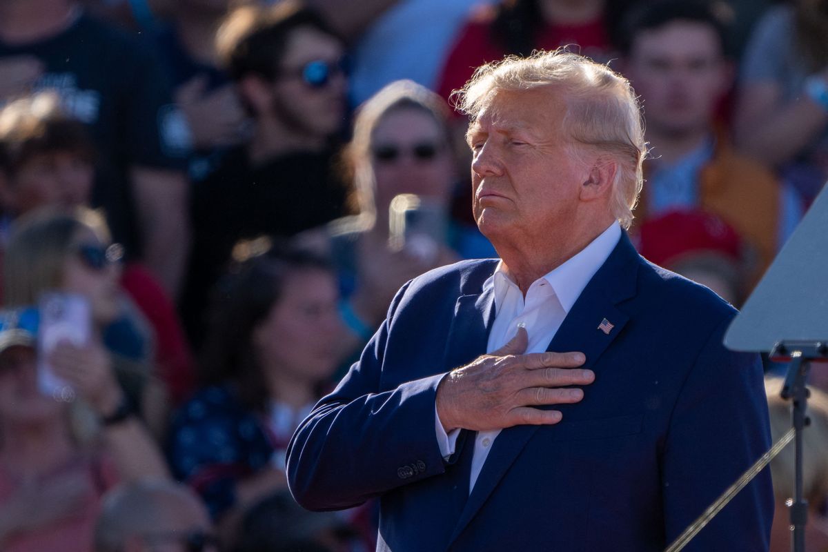 A white man wearing a blue suit with an American flag pinned on it stands with his hand on his hear. Many people are photographed behind him.