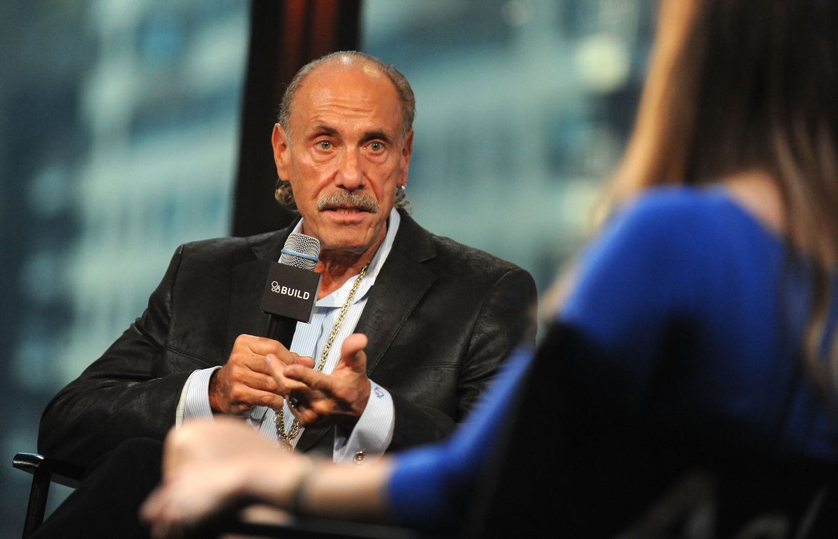 A white man wearing a black suit holds a microphone as he looks at a person facing him.