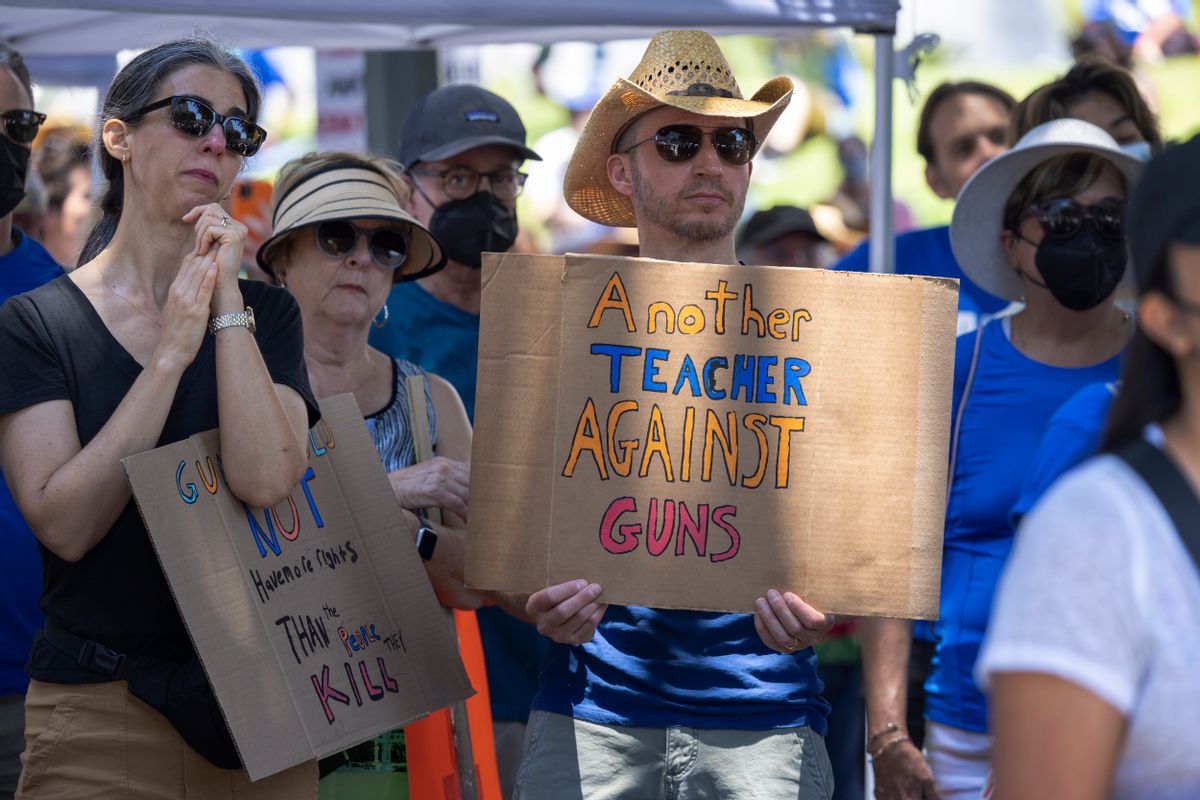 People stand gathered in a crowd. One wearing a cowboy hat says, "Another Teacher Against Guns."