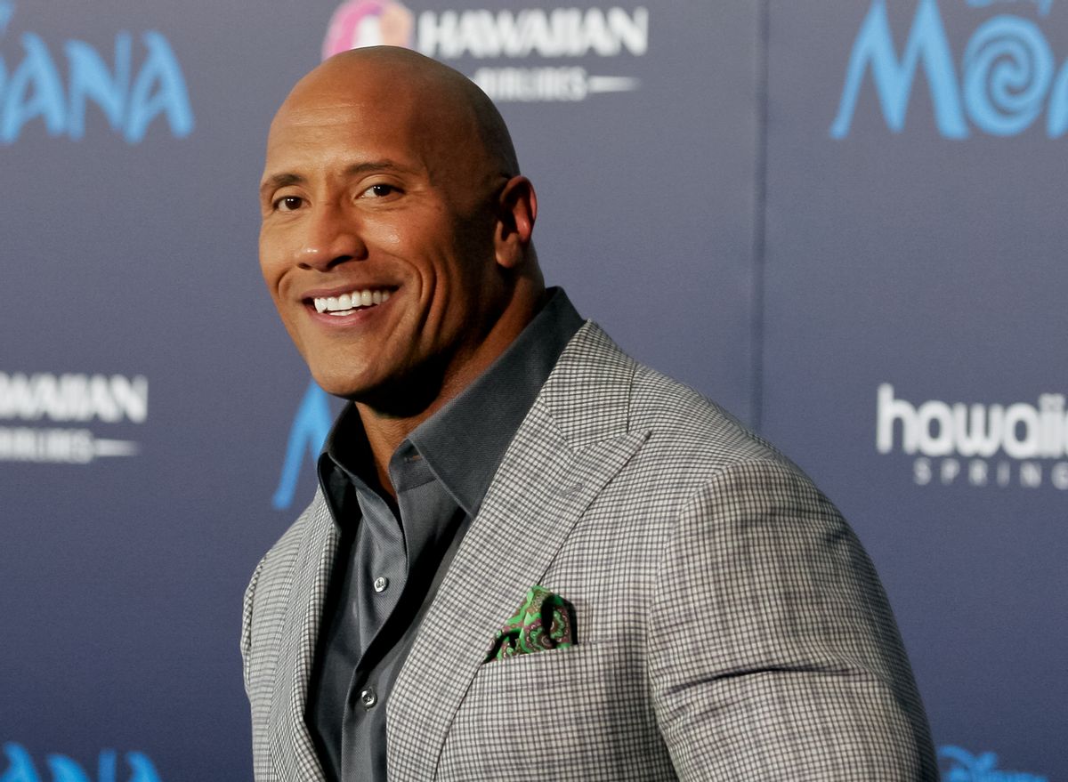 A Black/Samoan man in a gray suit smiles looking towards the camera.