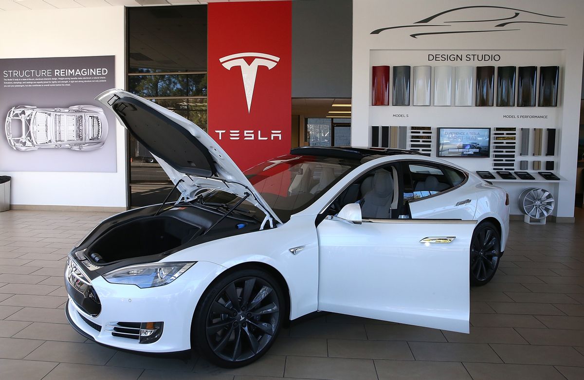 A white car with the driver's door and hood open is in front of a sign that says Tesla.