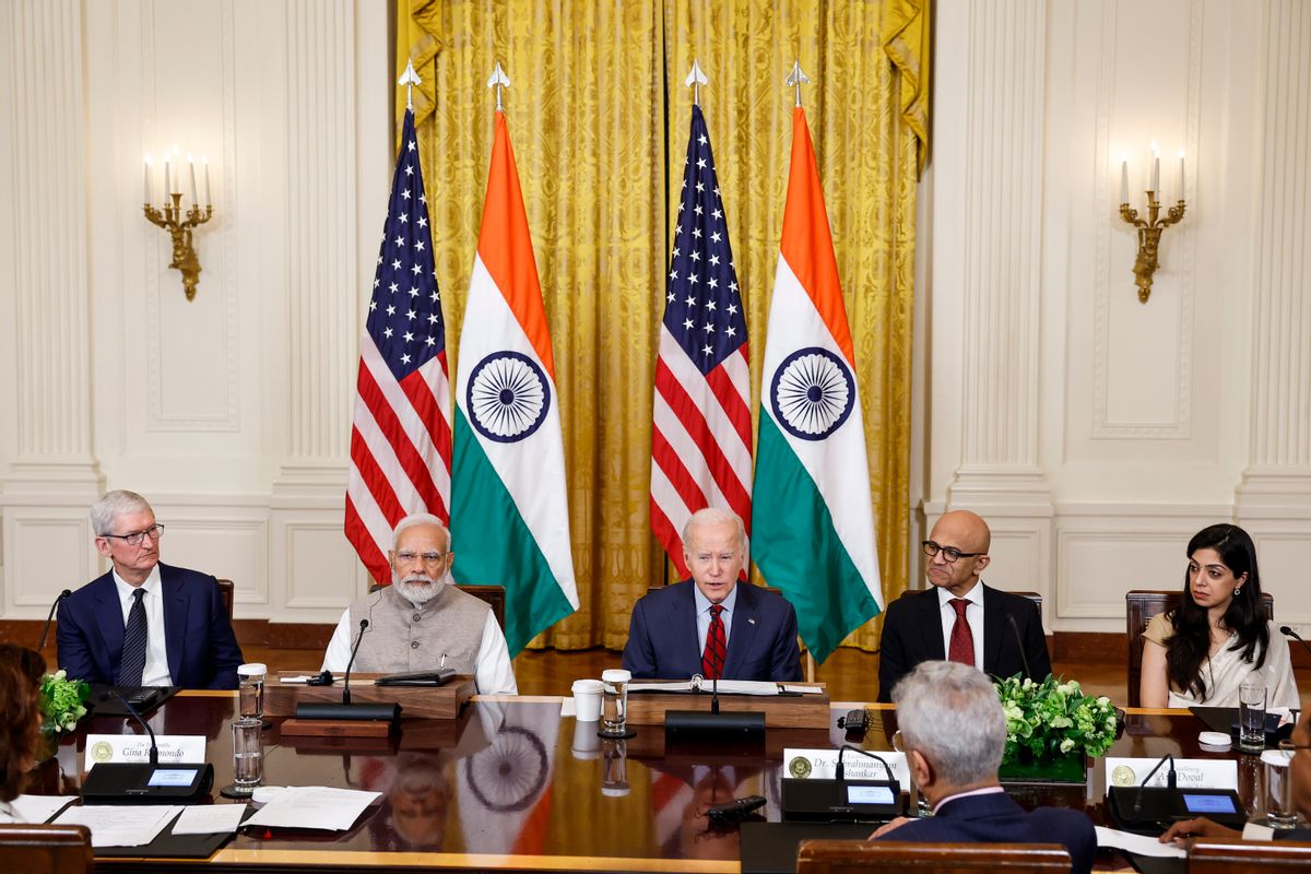 A white man sits in the middle of a table, surrounded by people on either side of him. In the background are two flags with red and white stripes and white stripes on a blue square, as well as two flags with orange, white and green stripes. On the white stripe of the flag is a spoked wheel.