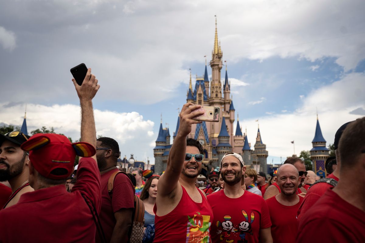 Two men wearing red shirts pose for a selfie surrounded by a crowd of people. Cinderella's Castle can be seen in the background.