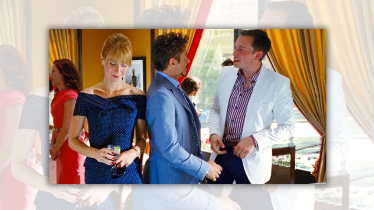 A man in a blue suit looks at a man wearing a white suit jacket and red and white striped button down. A woman with blonde hair looks down on the man in the blue suit's left.