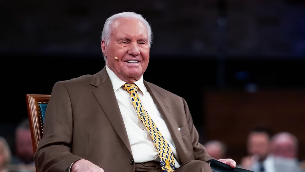 A white man in a brown suit smiles while sitting in a chair. A crowd of people sit behind him.