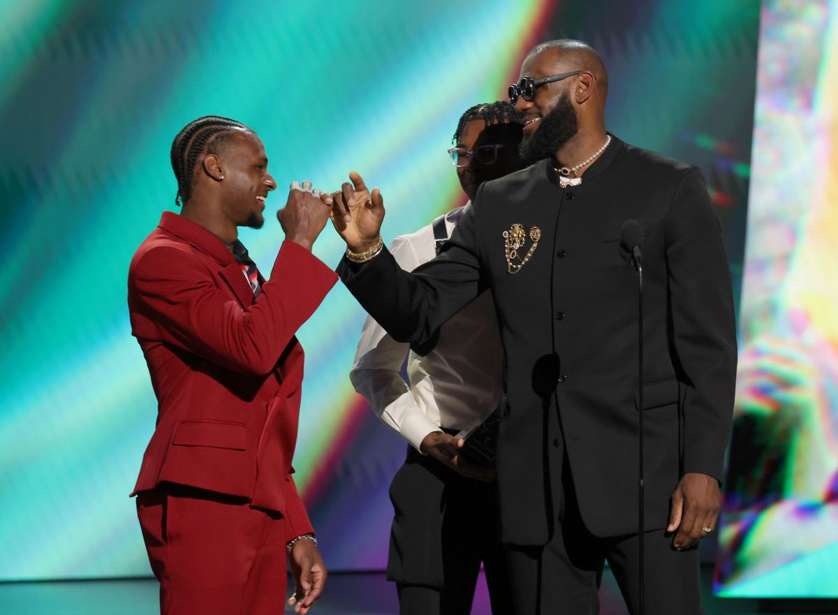 Two Black men smile at each other while linking pinkies. The one on the left is wearing a red suit, while the one on the right is wearing a black suit. A Black boy can be seen faintly behind the Black man on the right.