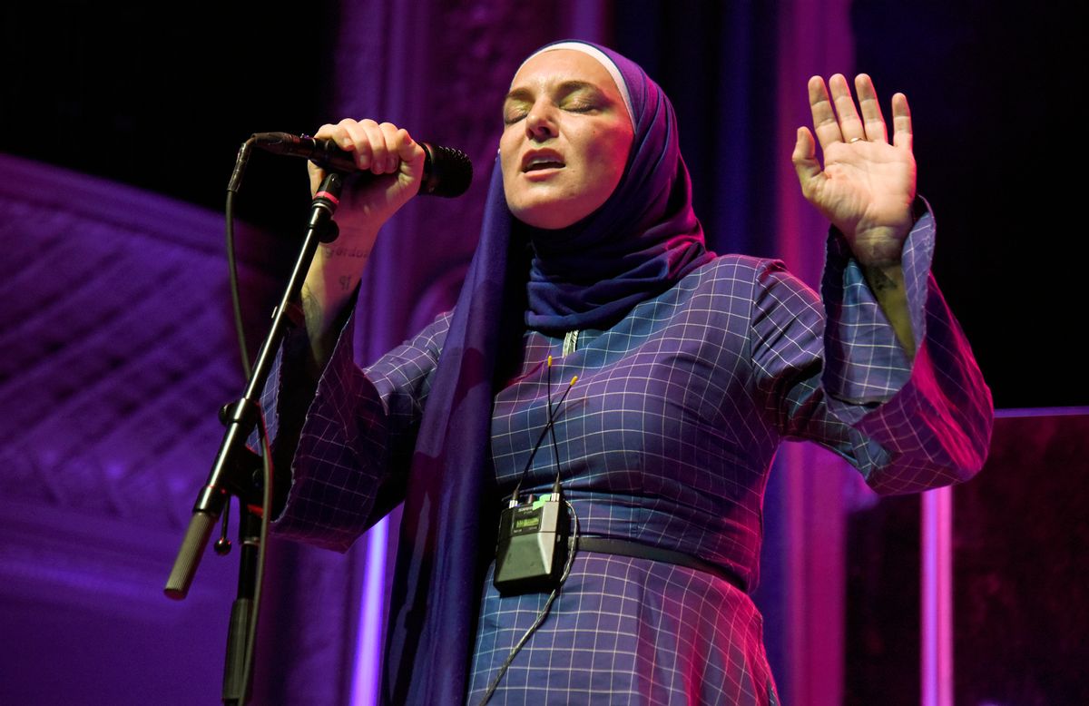 A white woman in a purple headscarf and dress holds a microphone in her left hand. Her right is held up, palm out. Her eyes are closed as she sings. 