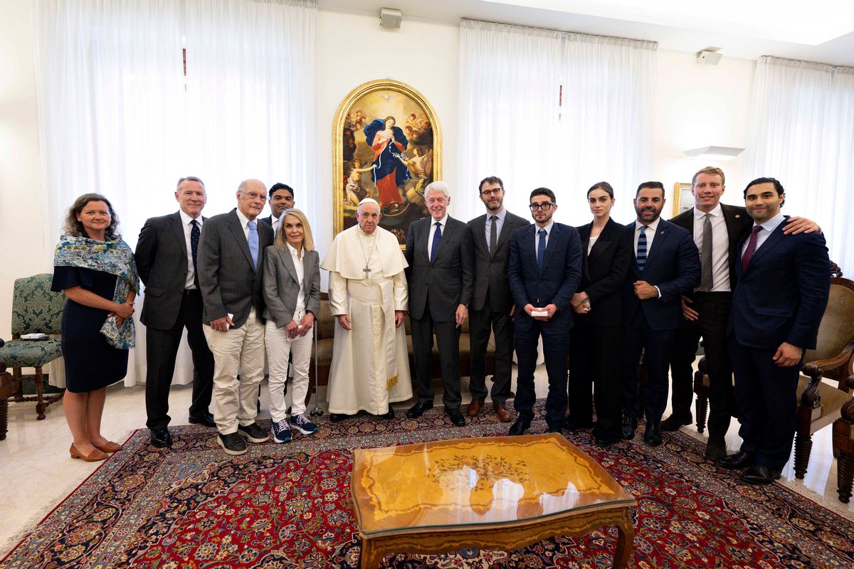 Several people gather together in a room to take a photo together. Many on the right are wearing black suits, while on the middle left a man wears all white with a cross around his neck.