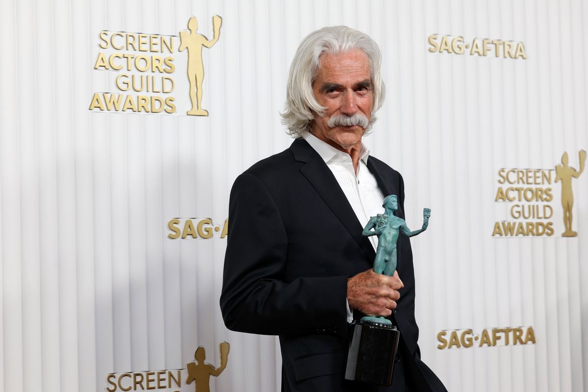 A white man with white hair and mustache wearing a black suit holds an award. 