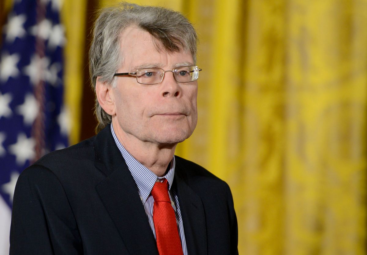 A white man wearing a suit and glasses is standing in front of a yellow backdrop. You can see the American flag behind him on his left side.