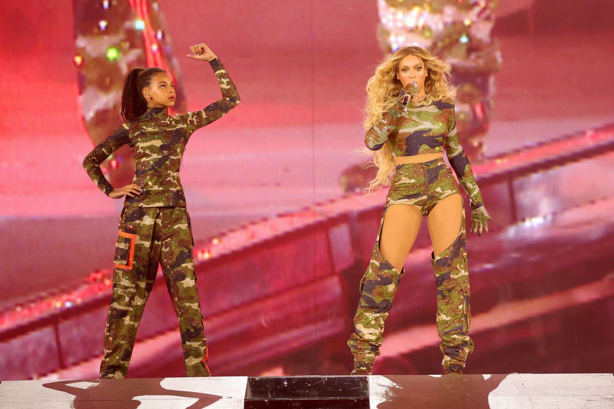 A Black girl wearing a camo outfit flexes her right bicep and looks up as a Black woman next to her (also wearing camo) sings.