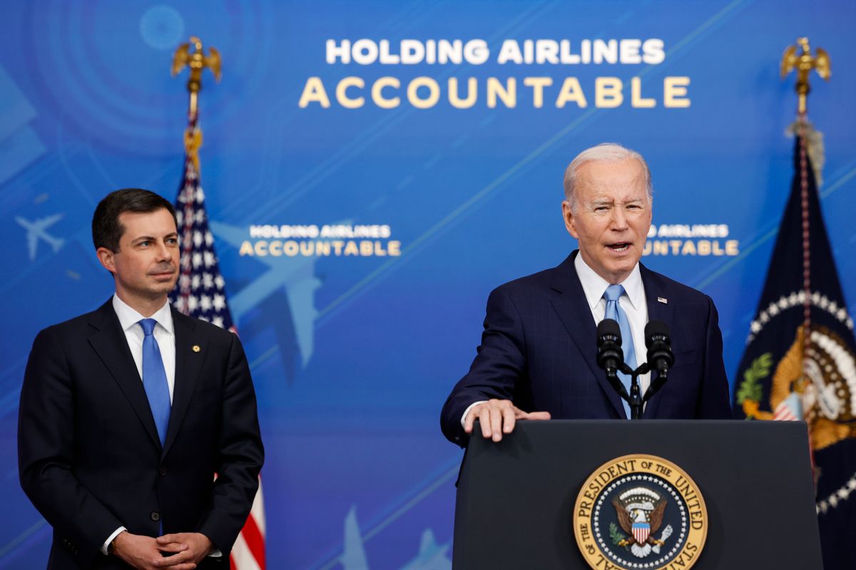 A white man wearing a black suit looks and listens to a white man standing at a podium that says President of the United States. A blue backdrop behind the two men says "Holding Airlines Accountable."
