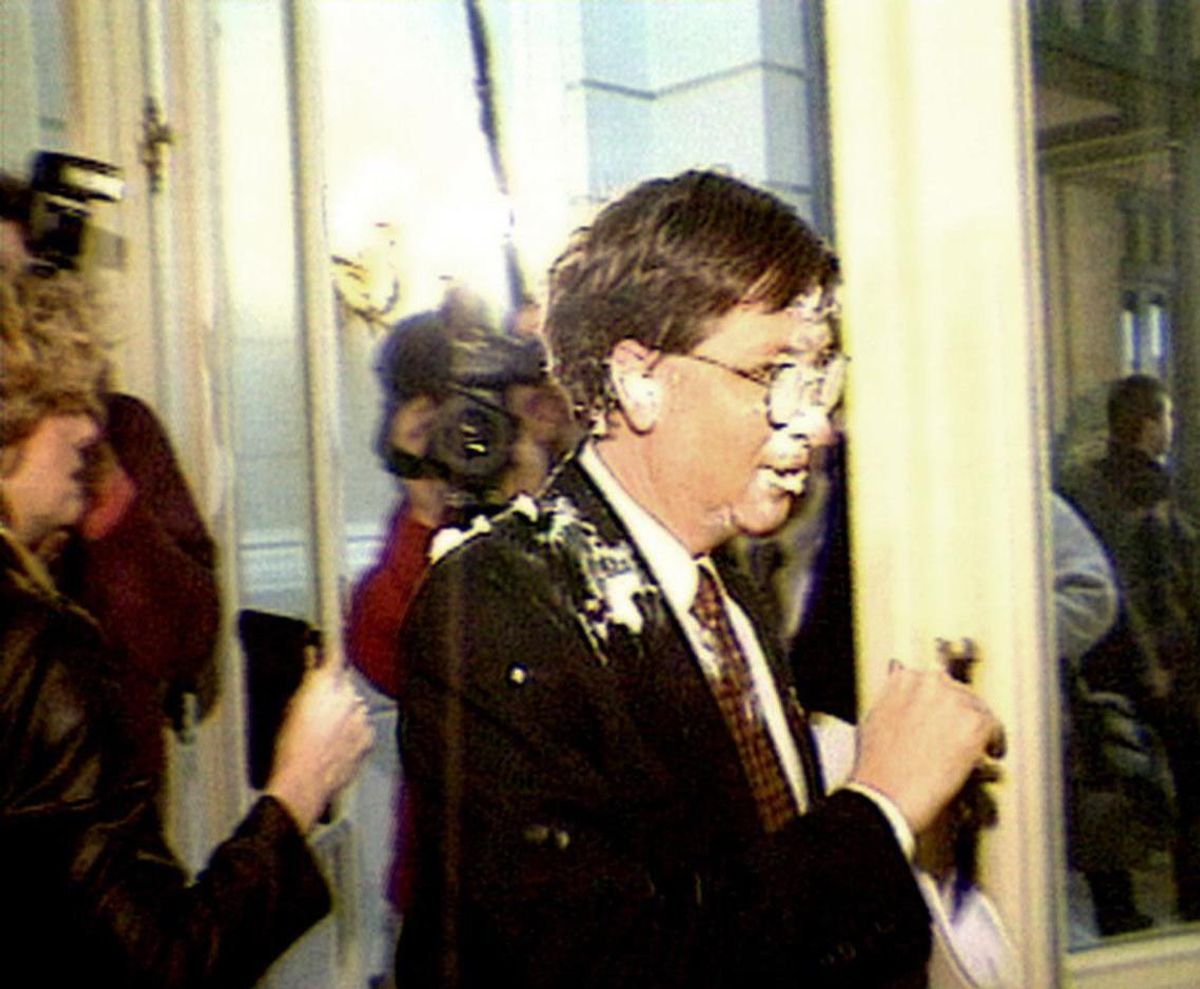 A white man in a black suit has white cream on his face, glasses, and suit. He looks shocked.