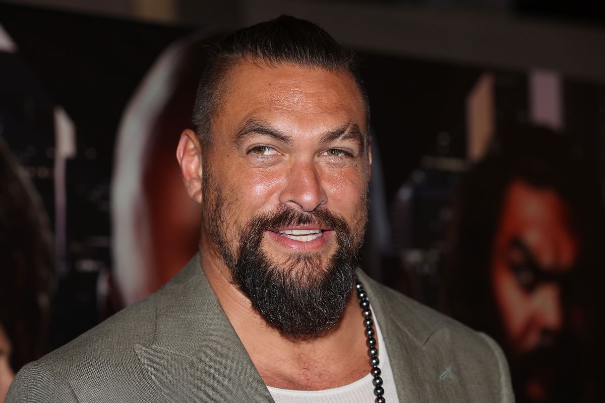 A Polynesian man with a beard wearing a gray suit is photographed smiling.