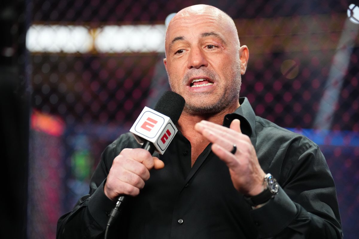 A white man is wearing a black button up and holding a microphone with red E's on each side.