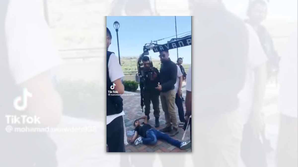 A Palestinian boy lies on the ground. People stand around him . You can see a film camera above him.