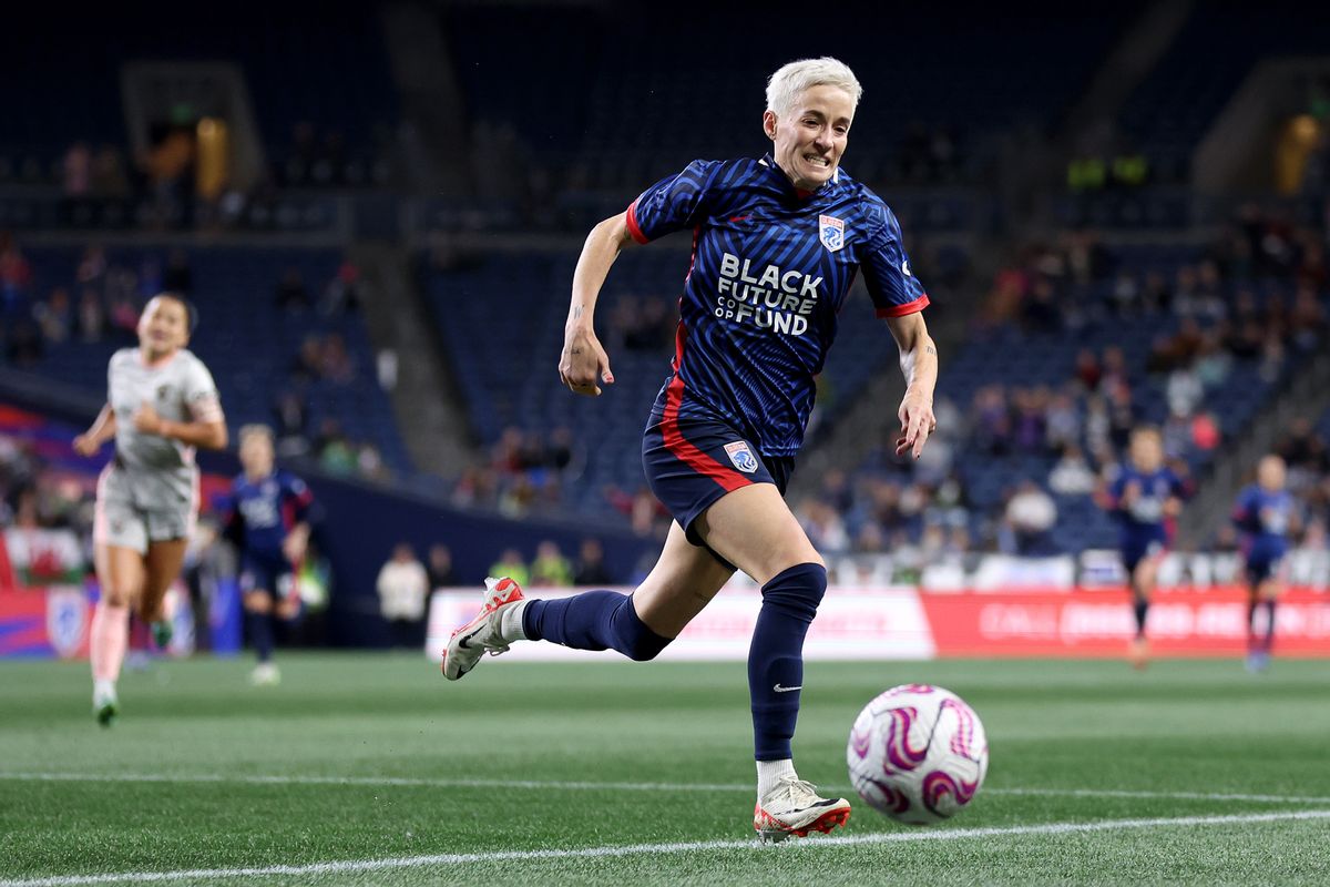 A white woman runs with a ball in front of her on a soccer field. Behind her run several other people. There are people sitting on the stands watching the match.