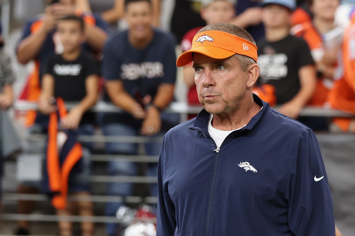 A white man wearing an orange visor and blue jacket that both have white horse logos on them stands on a football field.