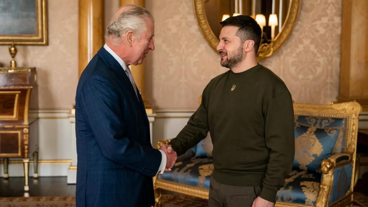 Two white men shake hands. THe one on the left is wearing a deep blue suit, while the one on the right is wearing a long-sleeve olive shirt.