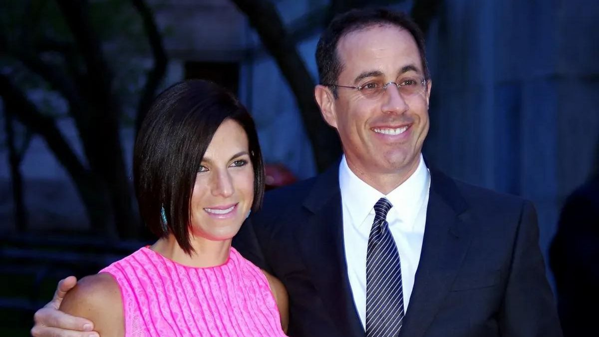 A white man stands next to a white woman, with his arm wrapped around her. Both are smiling.