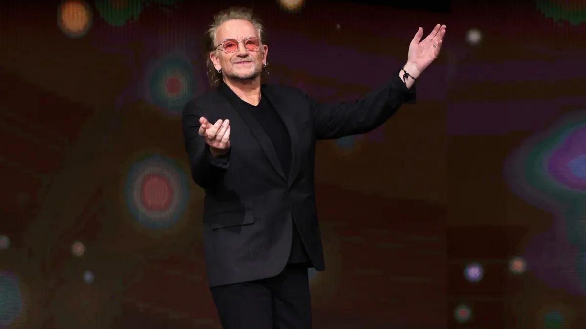 A white man wearing a black suit holds his hands open while facing forward.