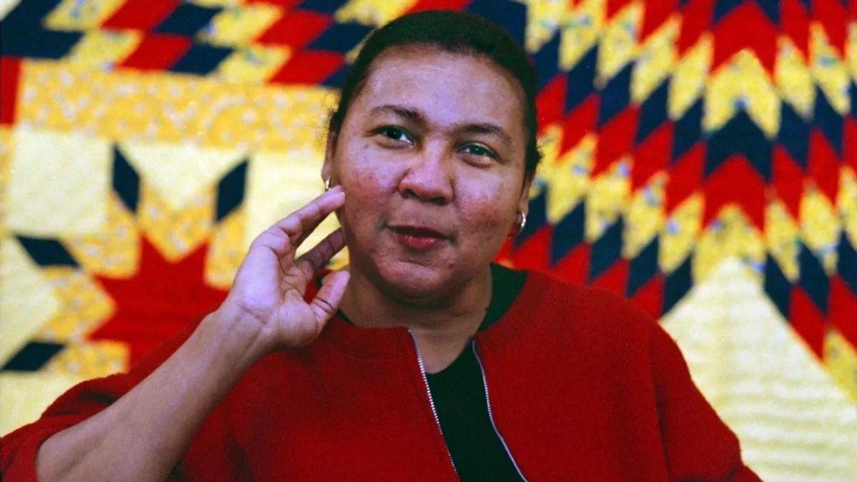 A Black woman wearing a red jacket holds her left arm to her face.