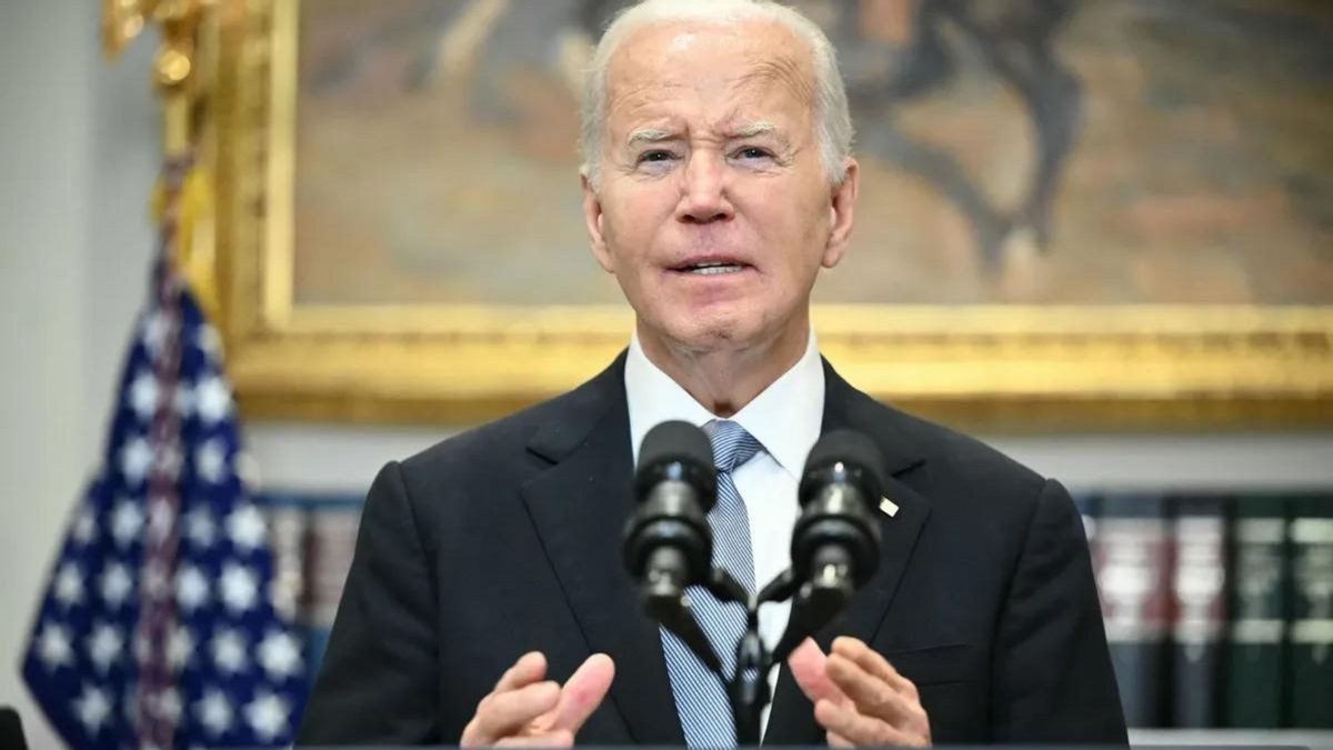 A white man wearing a black suit stands behind a podium that has a microphone on it.