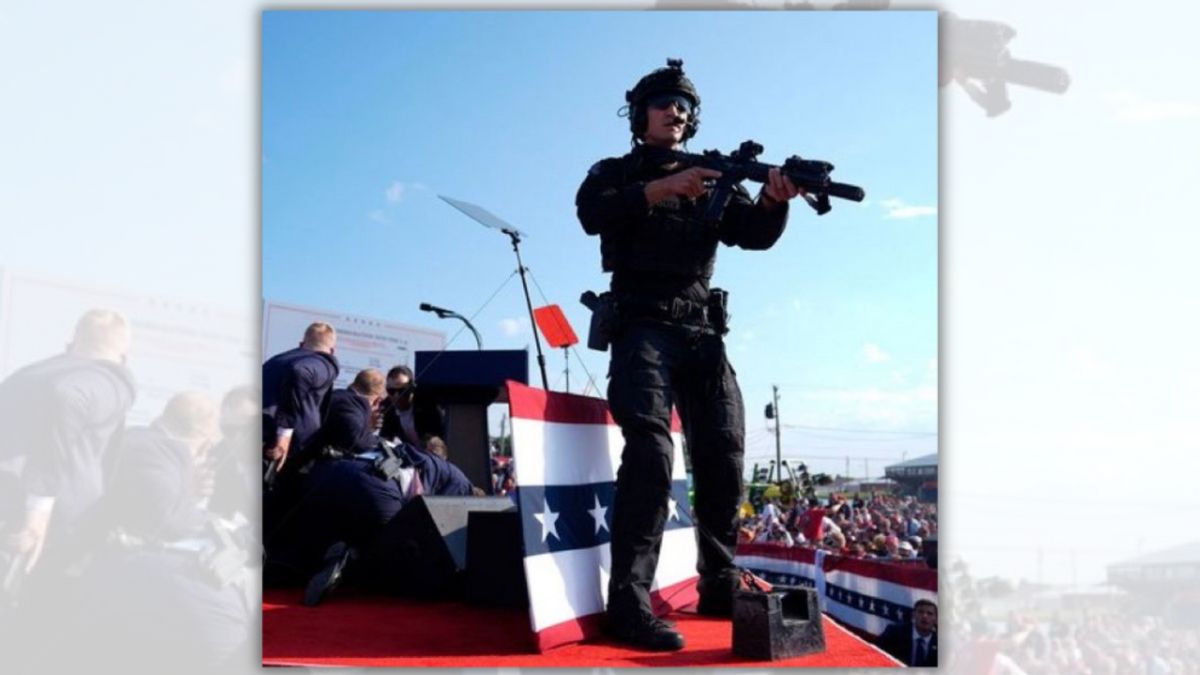 A person holding a rifle and wearing a black uniform is standing on a platform. You can see a crowd of people distantly to the side, as well as other people huddled near a podium.