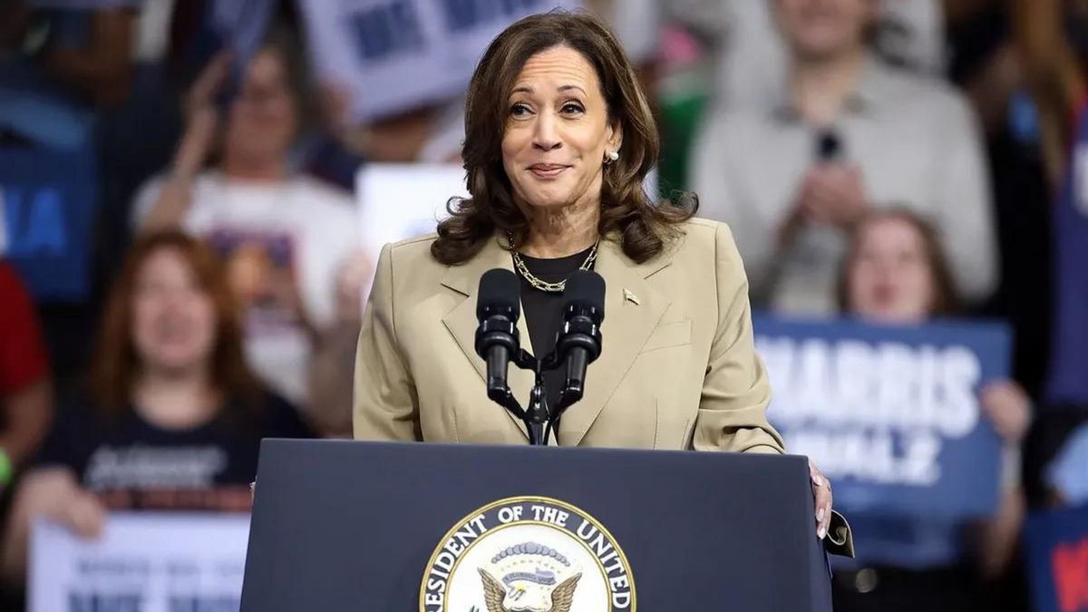 A Black Asian woman wearing a gray suit stands at a podium with a microphone on it. Behind her, you can see people standing that were holding signs of support.