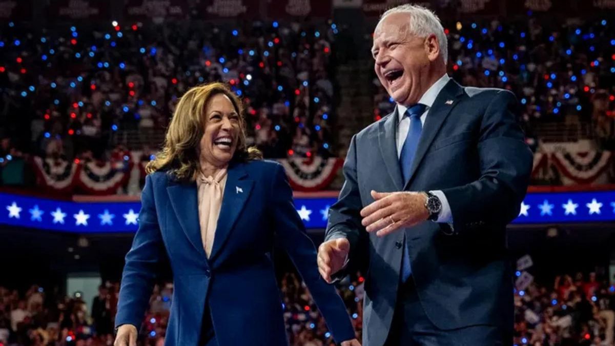 A Black Asian woman in a blue suit stands next to a white man wearing a gray blue suit. Both look happy and are standing in front of a large crowd.