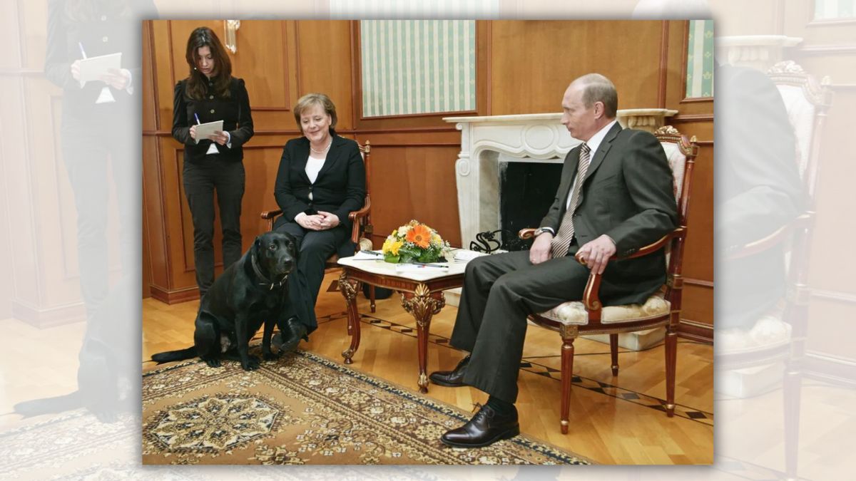A black lab sits next to a white woman with her hands crossed over her lap. A white man sits in another chair next to her. Another person stands on the other side of the woman.