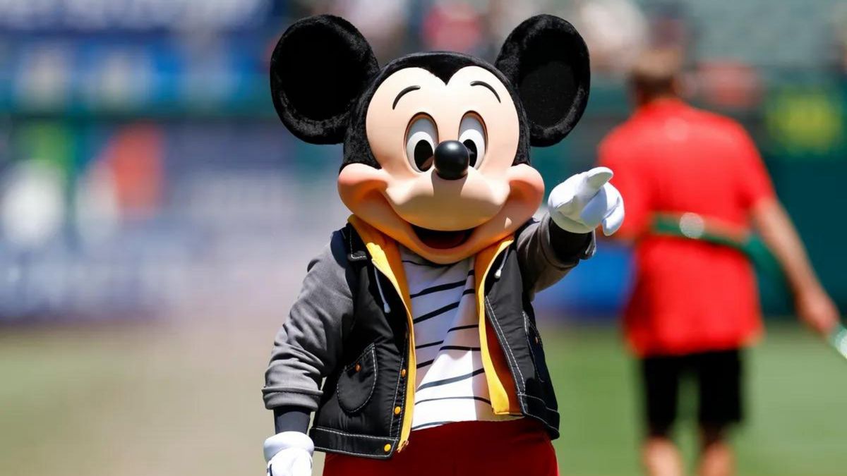 A mouse mascot wearing a black jacket, white and black striped shirt and red pants points to the right.