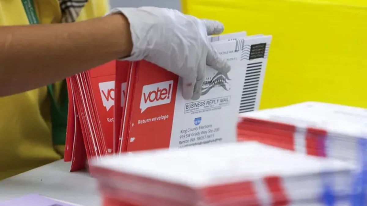 A Black person wearing a glove holds envelopes that says vote! on them next to stacks of envelopes.