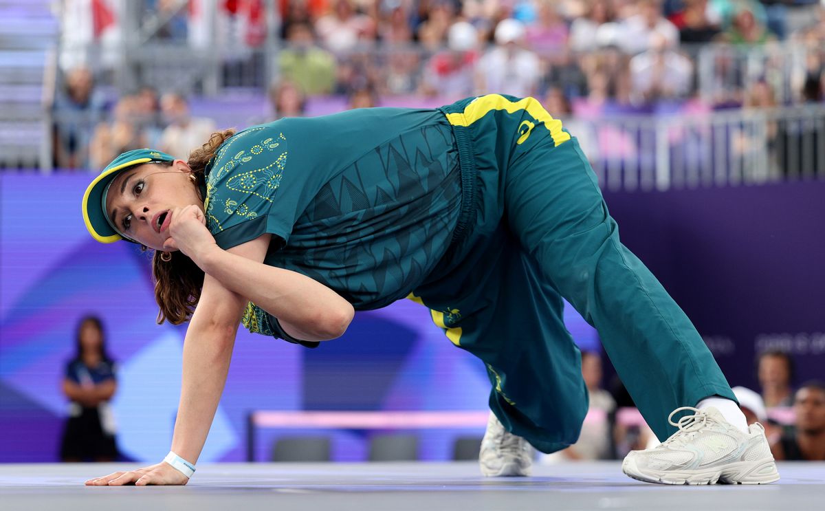 A white woman wearing a green and yellow shirt, pants and hat with white tennis shoes supports herself on one arm. Her other arm is wrapped around it.