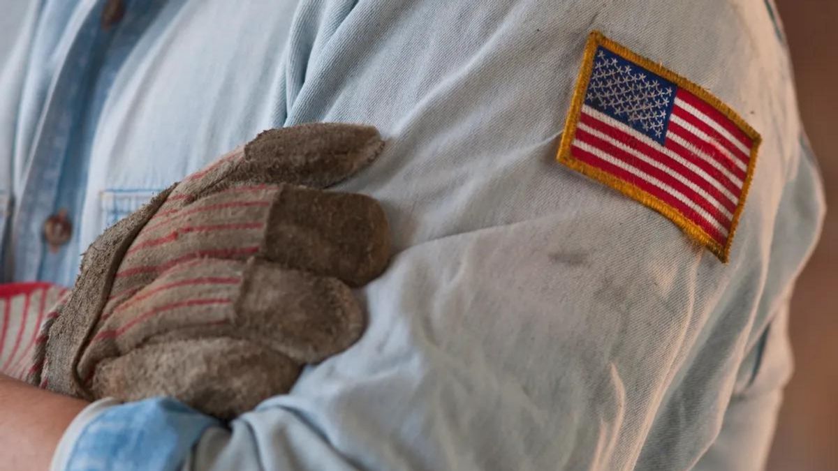 A person wearing a blue button down shirt with an American flag patch on it has their arms crossed. One hand is covered in a brown glove.