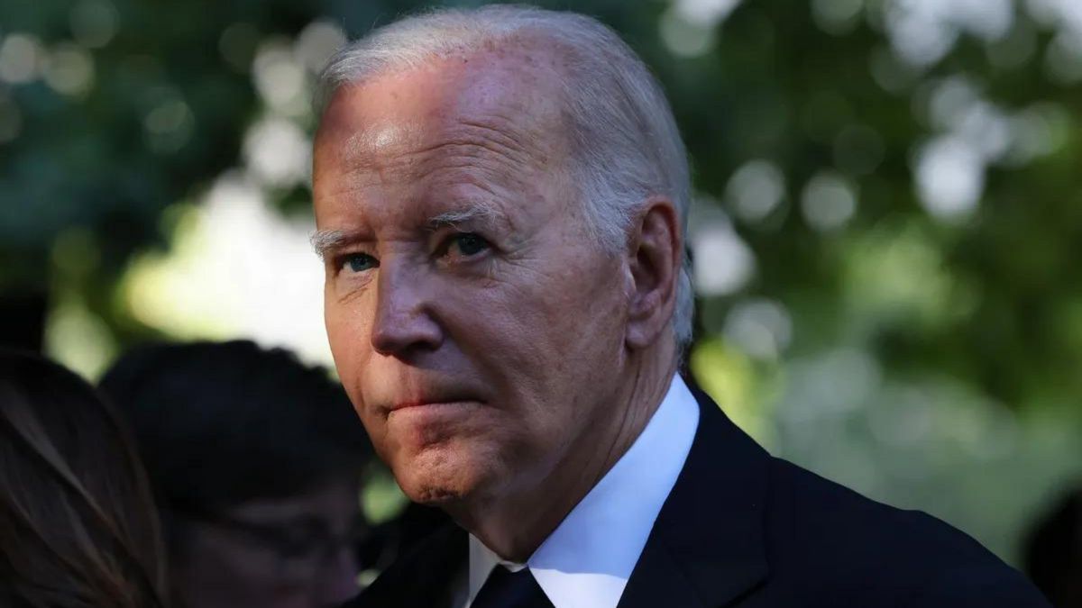 A white man wearing a black suit looks towards the camera. His face is partly shrouded by shadows.