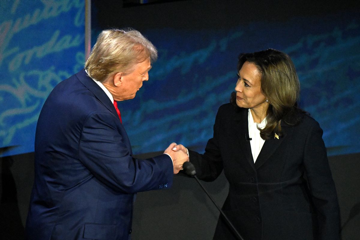 A white man wearing a blue suit shakes hands with a Black Asian woman wearing a black suit.