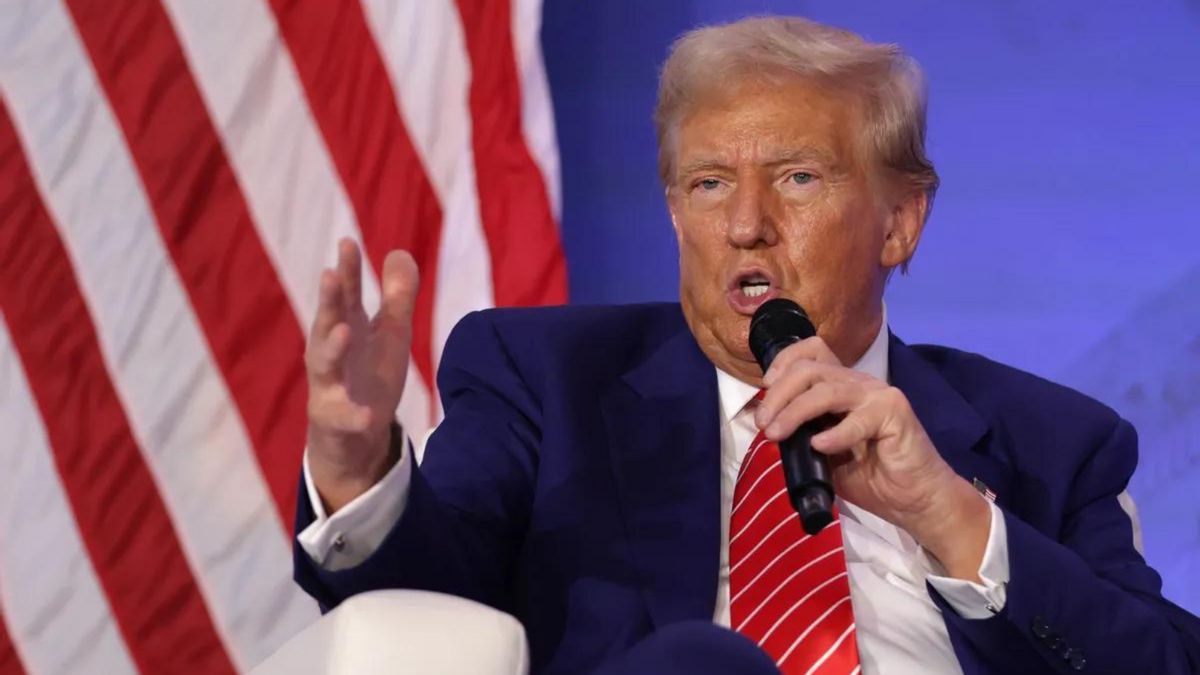 A white man wearing a blue suit speaks into a microphone while sitting down. A red and white striped flag can be seen behind him.