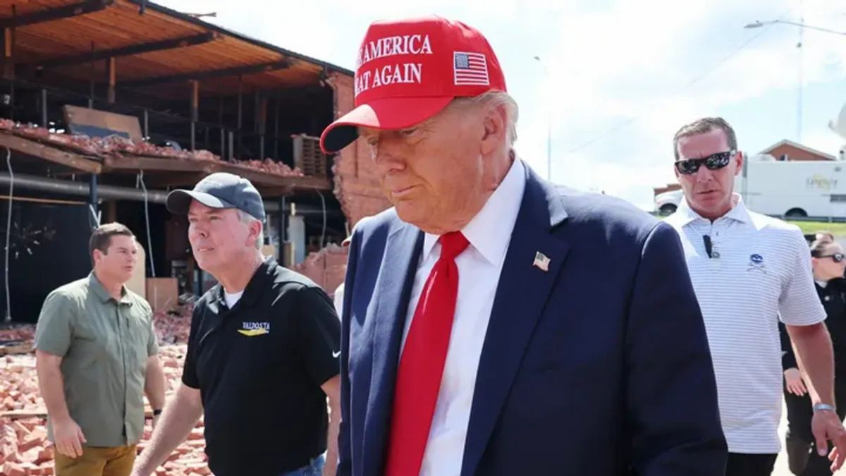 A white man wearing a red MAKE AMERICA GREAT AGAIN hat and blue suit looks down. The American flag can be seen on the side of the hat. Several people can be seen on either side of him.