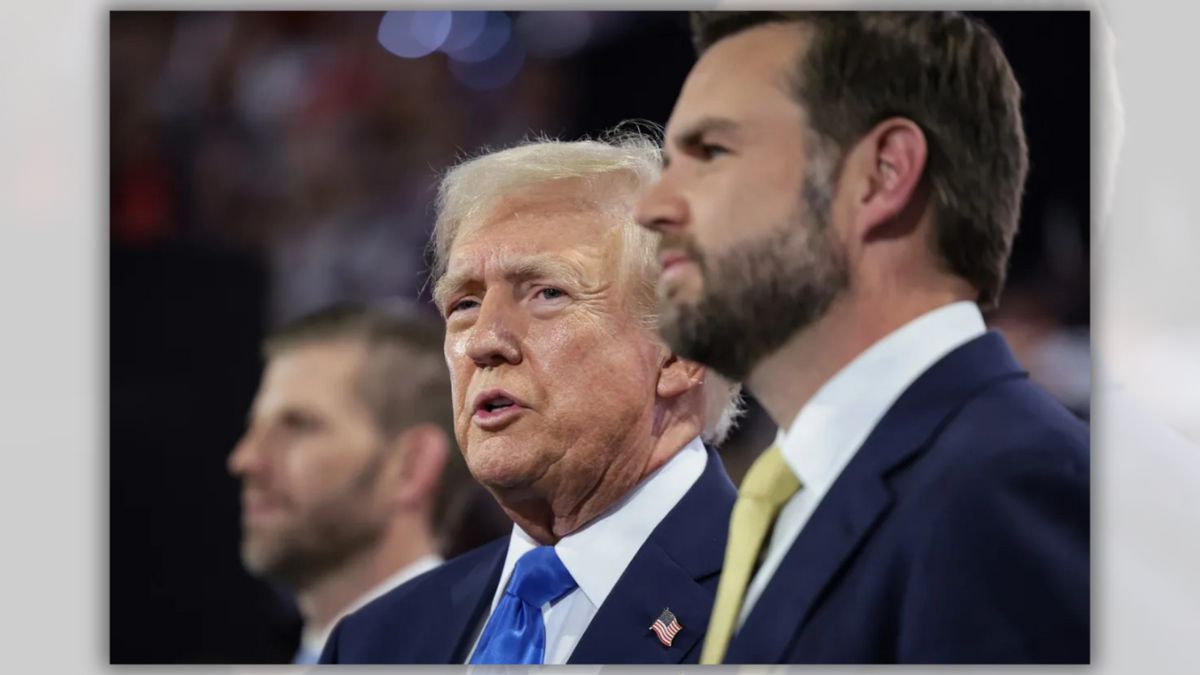 A white man wearing a blue suit stands next to another white man wearing a blue suit. A white person can be seen in the background of the image.