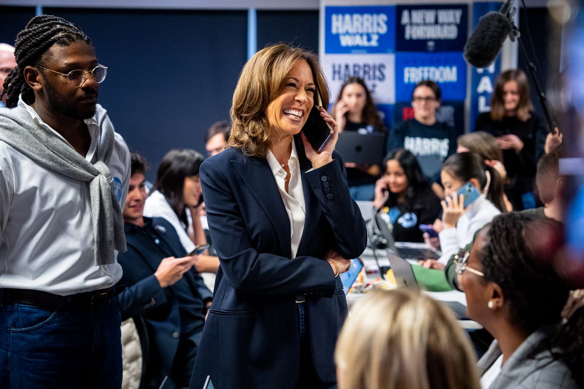 A Black South Asian woman wearing a blue suit smiles while holding a phone next to her ear. People surrounding her all look her way.