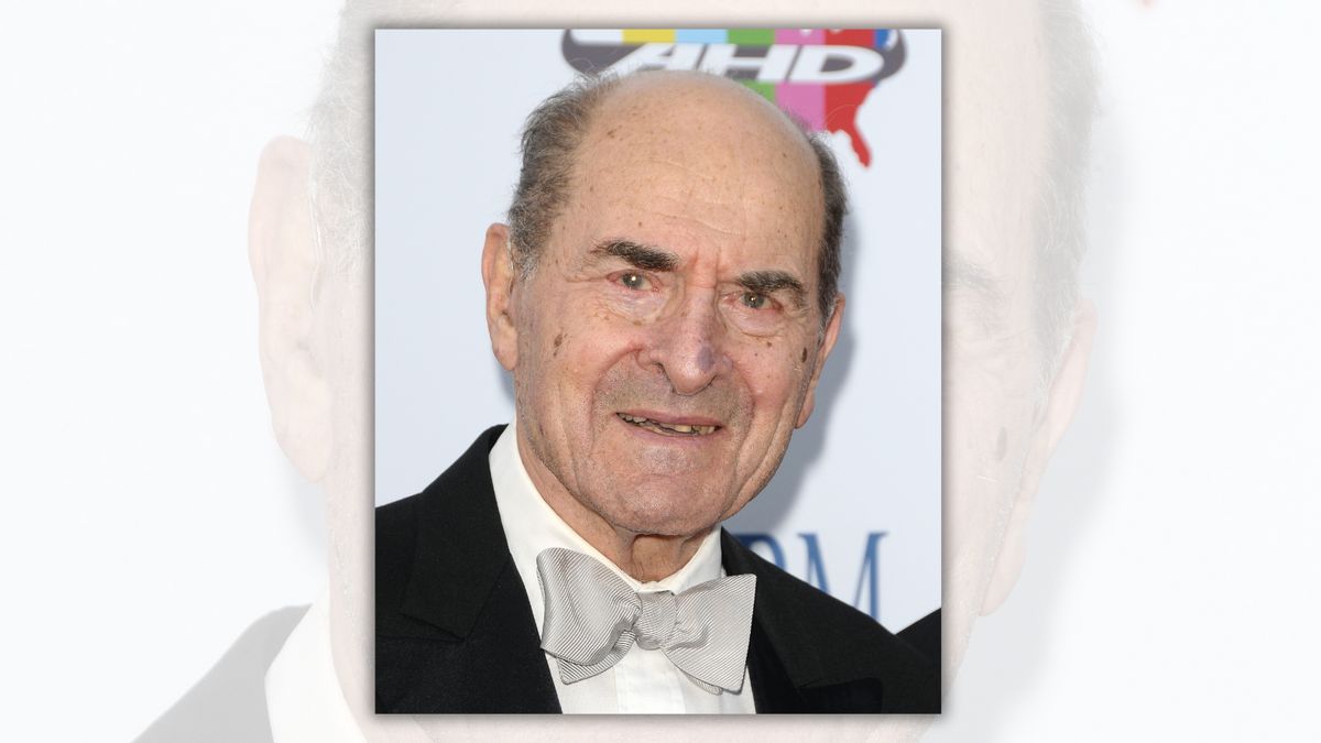 A white man wearing a black and white suit looks toward the camera.