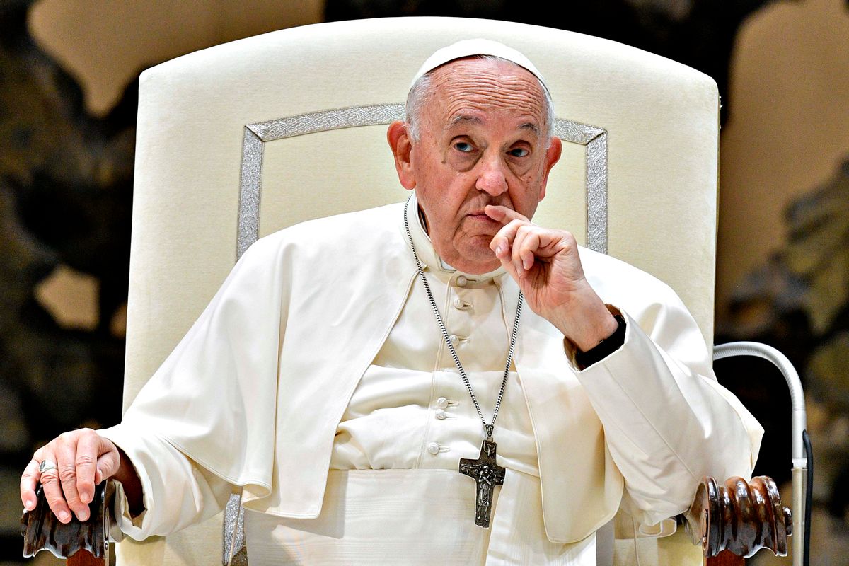 Pope Francis in traditional white papal clothing sitting on a big white chair, with his finger held to his mouth in thought.