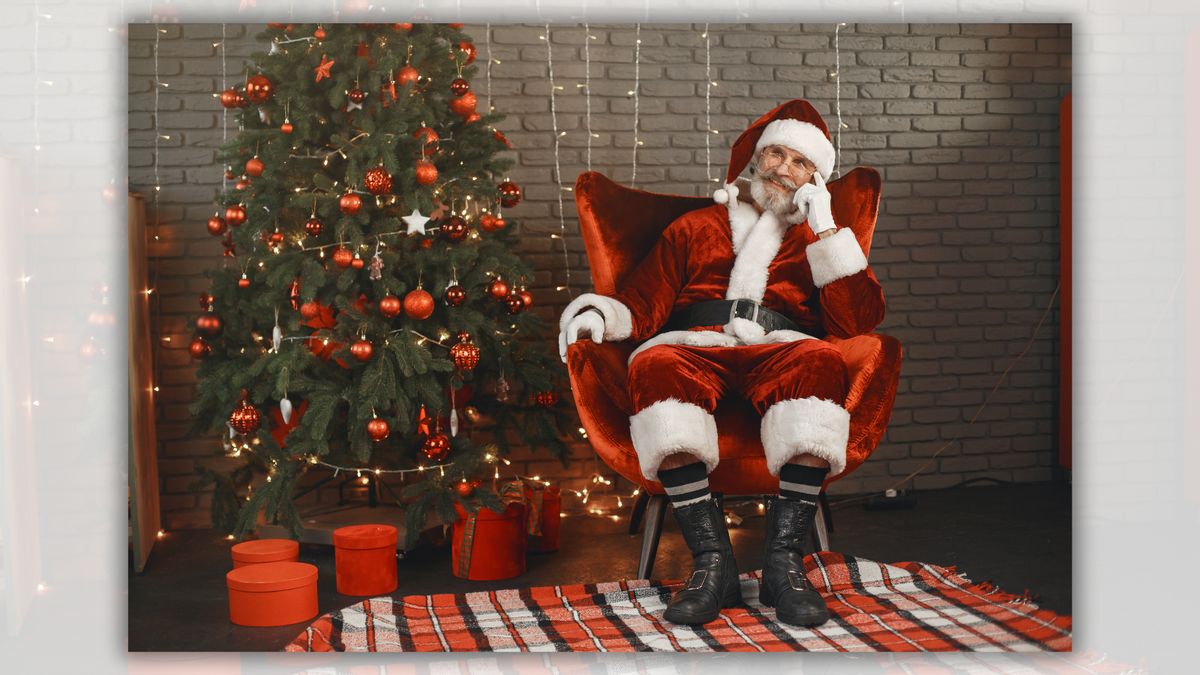 A white man sitting in a red chair wearing a red hat and sui is pictured next to a tree decorated with ornaments with presents underneath it.