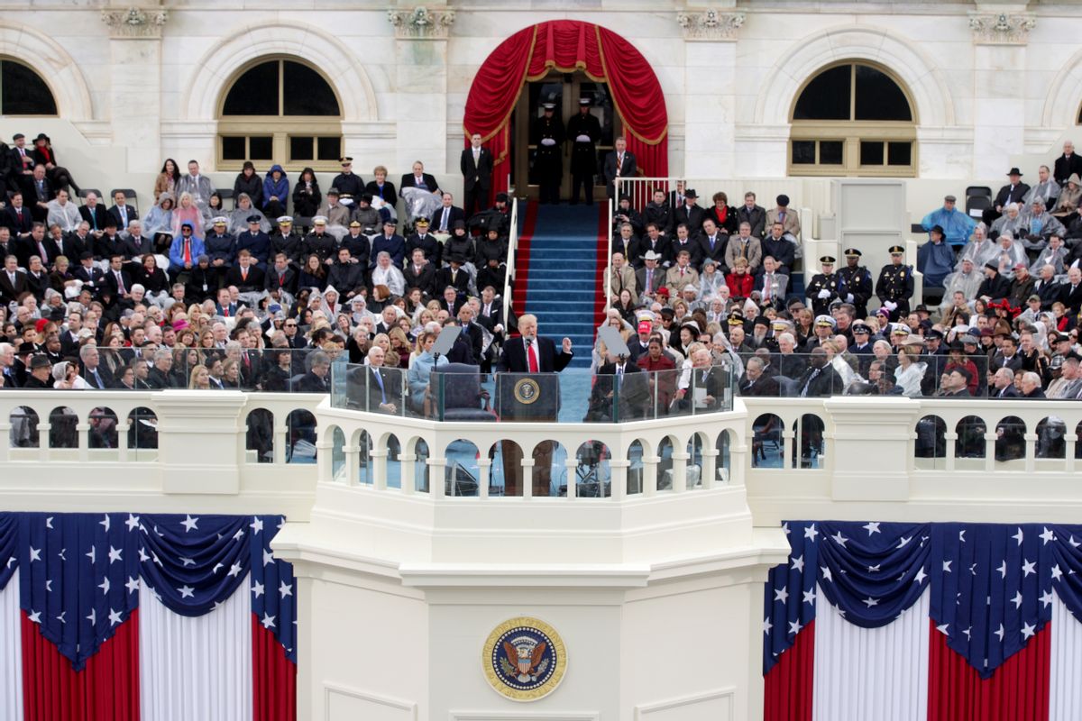 A wide shot of U.S. President Donald Trump's inauguration. Trump is standing at a podium in the center of the image, surrounded by a crowd of people sitting and watching him. The seal of the United States and US flags decorate the platform that he is standing on top of. The building in the backdrop is white marble. 
